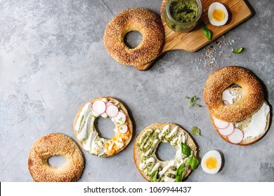 Variety Of Homemade Bagels With Sesame Seeds, Cream Cheese, Pesto Sauce, Eggs, Radish, Herbs Served On Crumpled Paper With Ingredients Above Over Grey Texture Background. Top View, Space.