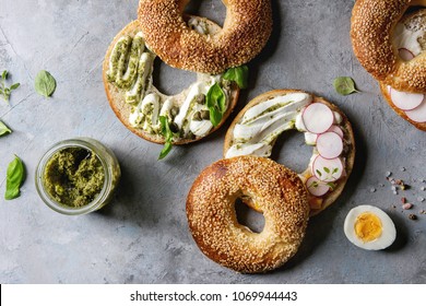 Variety Of Homemade Bagels With Sesame Seeds, Cream Cheese, Pesto Sauce, Eggs, Radish, Herbs Served On Crumpled Paper With Ingredients Above Over Grey Texture Background. Top View, Space.