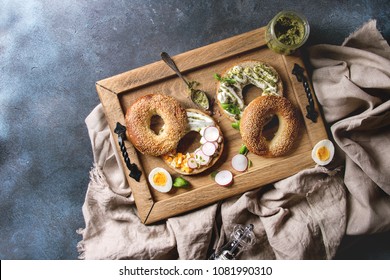 Variety Of Homemade Bagels With Sesame Seeds, Cream Cheese, Pesto Sauce, Eggs, Radish, Herbs Served On Wooden Tray With Cloth And Ingredients Above Over Blue Texture Background. Top View, Space.