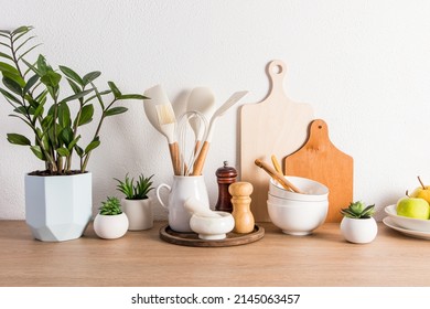 a variety of kitchen utensils, utensils, a potted flower and fruit on a plate on a wooden countertop against a white textured wall. front view