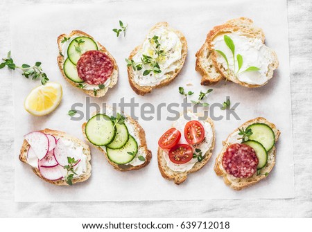 Variety of mini sandwiches with cream cheese, vegetables and salami. Sandwiches with cheese, cucumber, radish, tomatoes, salami, thyme, lemon zest on a light 
 background, top view. Flat lay   