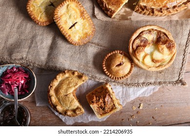 A Variety Of Pies Sat On A Hessian Background. Cheese Pie Cut In A Slice. Meat Pie, Steak Pie, Red Cabbage And Chutney. 