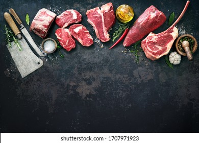 Variety of raw beef meat steaks for grilling with seasoning and utensils on dark rustic board