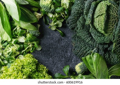 Variety of raw green vegetables salads, lettuce, bok choy, corn, broccoli, savoy cabbage as frame over black stone texture background. Top view, space for text