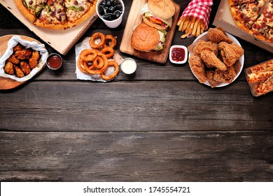 Variety Of Take Out And Fast Foods. Pizza, Hamburgers, Fried Chicken And Sides. Top Border. Above View On A Dark Wood Background With Copy Space.