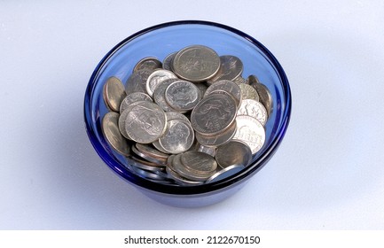 A Variety Of US Silver Coins In A Blue Glass Dish Set Upon A White Surface.