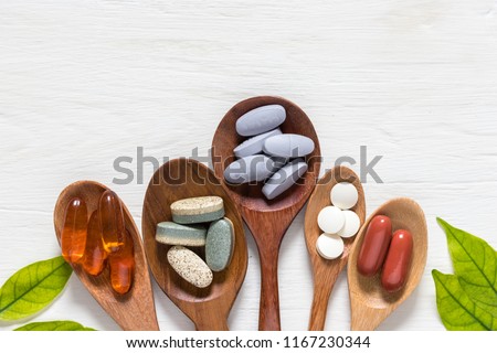 Variety of vitamin pills in wooden spoon on white background with green leaf, supplemental and healthcare product, flat lay surface
