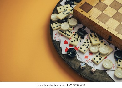 Various Board Games Chess Board, Playing Cards, Dominoes On A Old Wooden Table Orange Background.