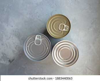 Various Canned Foods In Tin Cans On Concrete Grey Background. Non-perishable, Long Shelf Life Food For Survival In Emergency Conditions. Food Supplies Crisis Stock For Quarantine Isolation Period