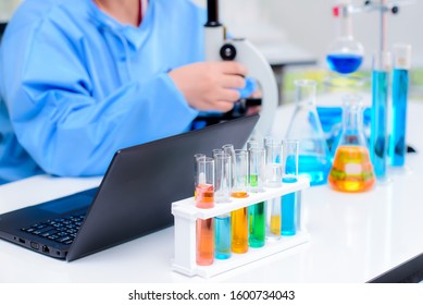 Various colored chemicals in a test tube placed on a scientist's desk in a lab. That has equipment to inspect and record results such as microscopes and laptops for data storage and analysis