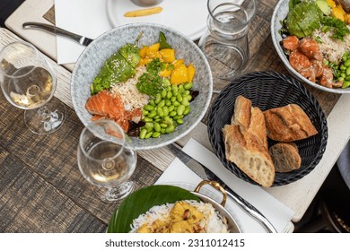 Various dishes and glasses with white wine lying on table of French restaurant. Salads with fish, avocado, rice, green beans and mango, bread arranged on plates on repast.