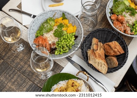Various dishes and glasses with white wine lying on table of French restaurant. Salads with fish, avocado, rice, green beans and mango, bread arranged on plates on repast.