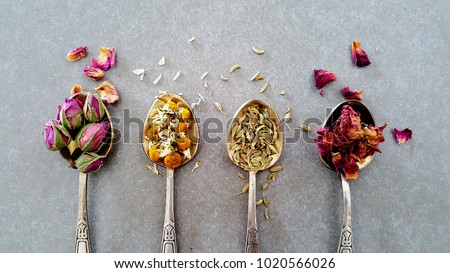 Various dry herbs and spices in vintage silver teaspoons. Flat lay of Indian Crumbled Roses, Fennel Seeds, Chamomile and Moroccan Roses on gray background. 