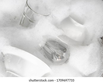 Various earthenware and glassware in soapy foam in a sink with soapy water
