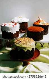 Various Easter cakes sit on cake stands during a holiday celebration in a bright room. The cakes have different toppings and frosting.