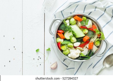 Various Fresh Vegetables In A Pot - Colorful Fresh Clear Spring Soup (vegetarian Bouillon Or Stock). Rural Kitchen Scenery From Above (top View). White Wooden Background - Layout With Free Text Space.