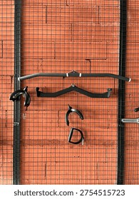 Various gym cable machine attachments and lat pulldown bars hanging on a black wire mesh rack against a red brick wall.