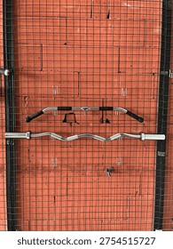 Various gym cable machine attachments and lat pulldown bars hanging on a black wire mesh rack against a red brick wall.