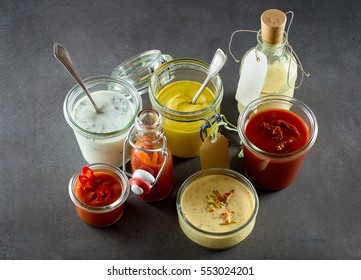 Various sandwich condiments in different sized glass containers and jars bunched together over gray background