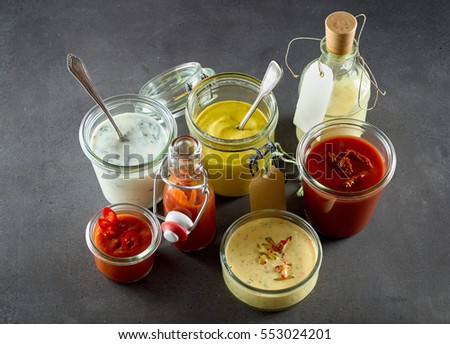 Various sandwich condiments in different sized glass containers and jars bunched together over gray background