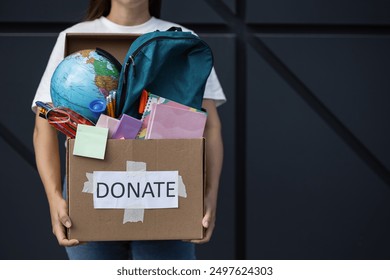 Various school supplies in a cardboard box in the hands of a volunteer on a geometric background with space for text. Conceptual donations. Back to school. Globe Backpack Notebook Pens - Powered by Shutterstock