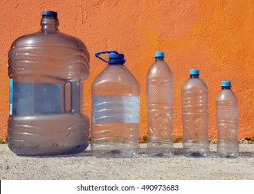 Various sized water bottles, arranged in descending order of volume from left to right, sits on a concrete ground in front of an orange wall.
