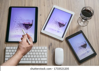 Various tablet computers, glass of red wine,  keyboard and mouse on the office desk. Drawing on the tablet computer.  Digital pencil in the artist's hand. Home office.