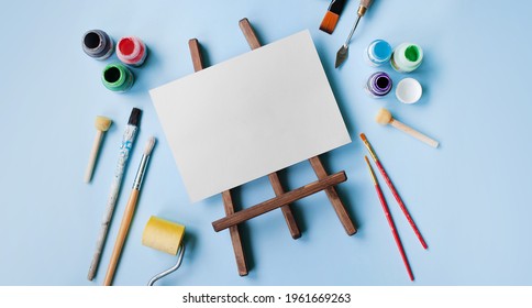 various tools for painting: brushes, spatula, roller, paints, blank on a wooden easel. flatley style on a blue background. copy space, mockup. art concept, learning to draw. baner style