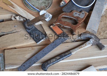Various traditional carpentry hand tools including claw hammer, try square ruler, hand saw, and adze on wooden boards in a workshop.