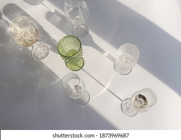 Various Transparent And Green Wine Glasses On A White Background And With Sun Shadows. Minimal Concept Of  Trendy, Dreamy, Romantic Celebration Photo With Wine Glasses And Their Shadows. 