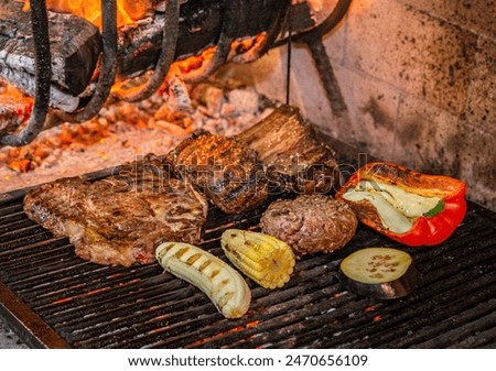 Various types of meat, pepper, corn, eggplant and banana being roasted in a typical Uruguayan grill called parrilla supplied with firewood and flames in a restaurant kitchen.