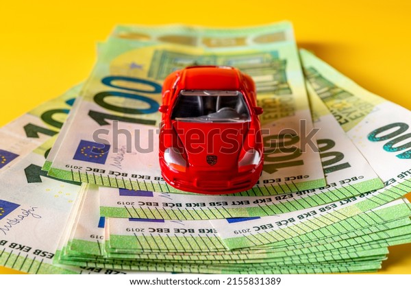 Varna,\
Bulgaria, May 10, 2022. Red Porsche miniature car model on a stack\
of 100 euro banknotes. Concepts of automobile insurance and auto\
loan. Car investment, money, wealth. Front\
view.