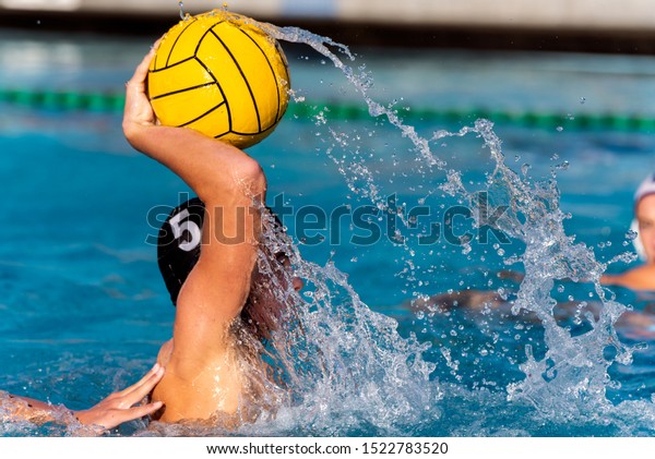 Varsity water polo player in black cap number 5 is\
bringing up ball for shot on goal with trail of splashing water\
showing his power.