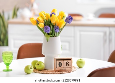 Vase with beautiful flowers, fresh apples, glass and cube calendar on dining table. International Women's Day celebration