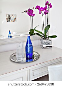 A Vase Of Pink Flowers Beside A Blue Sparkling Water Bottle On A White Table On A Hotel Bedroom