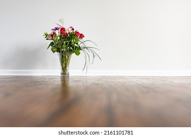 A Vase With A Rose Bouquet Placed On Hardwood Floor Next To A Wall With Baseboard In An Empty Domestic Room.