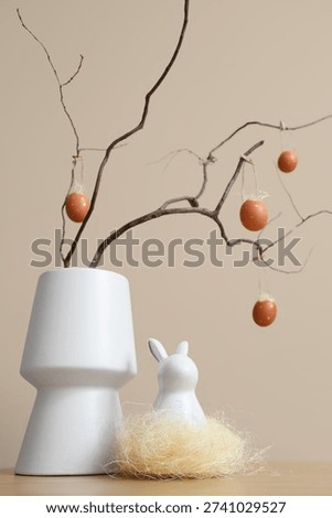 Vase with tree branches, Easter eggs and decorative rabbit on table near beige wall