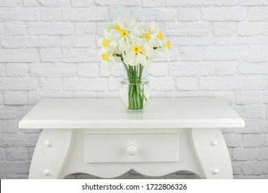 Vase Of White And Yellow Daffodil Flowers Displayed On A White End Table With A White Brick Wall Background