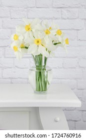 Vase Of White And Yellow Daffodil Flowers Displayed On A White End Table With A White Brick Wall Background