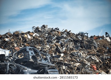 A vast accumulation of discarded vehicles and assorted metal debris forms a large mound against a backdrop of a partly cloudy sky