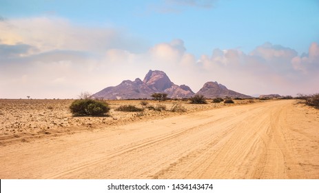 A vast African landscape scene, with a dirt road running through barren flat plains in the Namib Desert toward the huge granite peaks of Spitzkoppe, Namibia. - Powered by Shutterstock