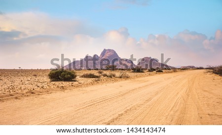 A vast African landscape scene, with a dirt road running through barren flat plains in the Namib Desert toward the huge granite peaks of Spitzkoppe, Namibia.