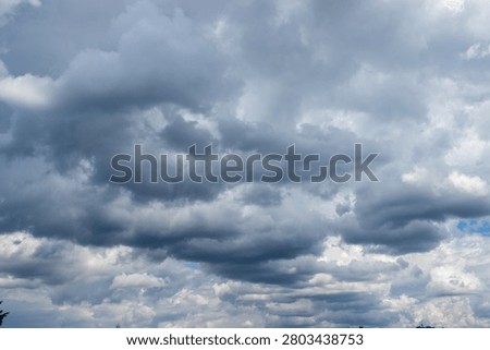 A vast dramatic sky dominated by a dynamic interplay of dark heavy grey clouds and bright white cumulus formations. Glimpses of clear blue sky peek through suggesting transitional or unsettled weather.