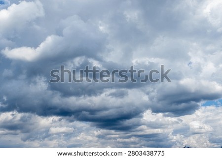 A vast dramatic sky dominated by a dynamic interplay of dark heavy grey clouds and bright white cumulus formations. Glimpses of clear blue sky peek through suggesting transitional or unsettled weather.
