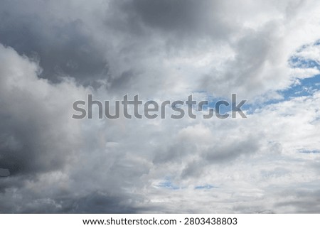A vast dramatic sky dominated by a dynamic interplay of dark heavy grey clouds and bright white cumulus formations. Glimpses of clear blue sky peek through suggesting transitional or unsettled weather.