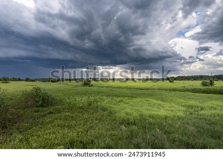 A vast, dramatic sky filled with dark storm clouds hangs over a lush, green field. The sunlight filters through the clouds, casting a soft glow on the landscape.