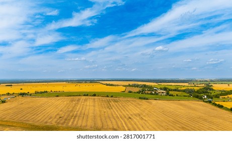 Vast fields under blue sky in summer. Golden fields stretch across the landscape with few trees and a house visible in the distance.