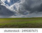 A vast green field stretches out beneath a sky filled with dramatic, dark clouds. The clouds are illuminated from behind, creating a sense of impending rain.
