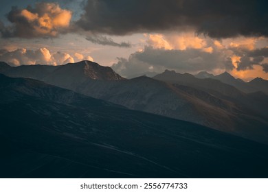 A vast mountain range lies under dark clouds, with rays of sunlight piercing through, casting brief, dramatic light across the jagged peaks and deep valleys below. - Powered by Shutterstock