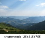 A vast mountain range stretches across the horizon, showcasing layers of green hills and blue skies during a sunny day. The scene is peaceful, highlighting natures beauty and tranquility.